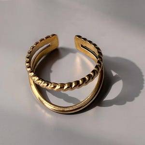 Op de afbeelding: Een verstelbare ring in goudkleur met een dubbele band. De bovenste band heeft een gedraaide touwtextuur, terwijl de onderste band glad is. De ring heeft een opening aan de bovenkant voor maatverstelling. Grijze achtergrond.