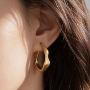 May include: A close-up of a gold-coloured earring with an irregular, wavy shape. The earring is a hoop style, with a smooth, polished finish. The earring is worn on the ear of a person with brown hair.