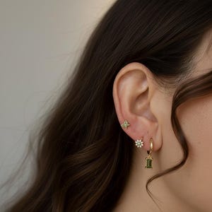 Könnte beinhalten: Goldene Ohrring-Set mit einem kleinen vierblättrigen Kleeblatt-Stecker, einem Gänseblümchen-Stecker und einem Creolen-Ohrring mit einem grünen, rechteckigen Edelstein. Die Ohrringe werden an einem Ohr getragen, mit dunklem Haar im Hintergrund.