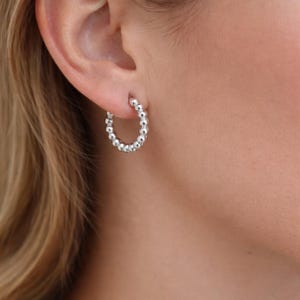 Könnte beinhalten: Silberne Creolen mit Perlenmuster. Die Ohrringe sind ein klassischer Stil mit einer Reihe kleiner, runder Perlen, die eine strukturierte Oberfläche bilden. Die Ohrringe sind an einem Ohr zu sehen.