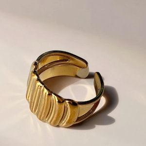 Könnte beinhalten: Ein goldfarbener, verstellbarer Ring mit geripptem Design auf der Vorderseite. Der Ring hat eine glatte, polierte Oberfläche und ist hinten offen zur Größenanpassung. Der Ring ist vor einem weißen Hintergrund platziert.
