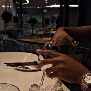 May include: A person pouring red wine into a clear wine glass at a restaurant. The scene includes a white table, a plate with silverware, and a bowl of food. A wristwatch is visible on the person's wrist.