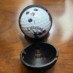 May include: A white Callaway golf ball with black markings and the number 1, held in a black, open, spherical case. The case features circular cutouts. The golf ball is the main subject.