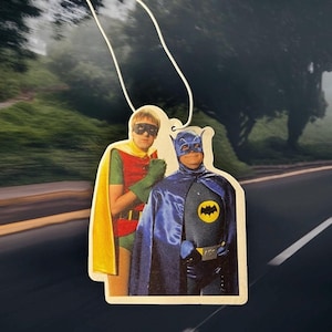May include: Air freshener depicting Batman and Robin in their iconic costumes. Batman is in a dark blue suit with a yellow bat emblem. Robin is in a red and green outfit with a yellow cape. The air freshener hangs from a white string.