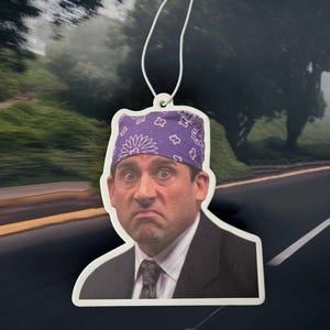 May include: Air freshener with a character wearing a purple bandana. The character has a surprised expression and is wearing a suit and tie. The air freshener is suspended from a white string.