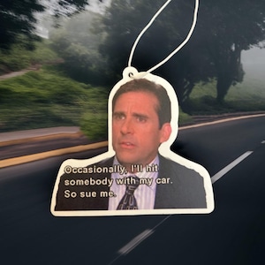 以下が含まれることがあります： 男性の写真と「Occasionally, I'll hit somebody with my car. So sue me.」という引用句がプリントされた白い芳香剤。吊り下げ用の白い紐が付いています。背景は、道路と木のぼやけた画像です。