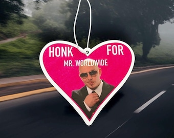 以下が含まれることがあります： ピンク色のハート型芳香剤。白い文字で「HONK FOR MR. WORLDWIDE」と書かれています。サングラスとスーツを着た男性の写真が特徴です。吊り下げ用の白い紐が付いています。