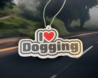 以下が含まれることがあります： 白いひもで吊るされた、赤いハートと「I ❤️ Dogging」の文字が入った白とグレーのデザインの芳香剤。道路と木のぼやけた背景に対して吊り下げられています。