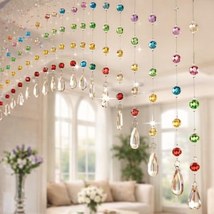 Puede incluir: Una cortina decorativa de cuentas con cuentas de cristal de colores y colgantes en forma de lágrima. Las cuentas están en un arcoíris de colores, incluyendo rojo, naranja, amarillo, verde, azul y morado. La cortina cuelga frente a una ventana.