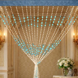 Puede incluir: Una cortina de cuentas con cuentas blancas y turquesas. La cortina cuelga de una cabecera de tela azul. Las cuentas están ensartadas en líneas verticales, creando un separador de ambientes o un tratamiento de ventana decorativo.