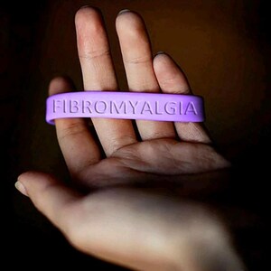 Może przedstawiać: Lawendowa silikonowa opaska na nadgarstek z wytłoczonym słowem FIBROMYALGIA wielkimi literami. Opaska jest trzymana w dłoni na ciemnym tle. Tekst jest łatwy do odczytania.