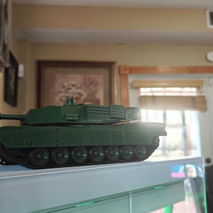 May include: A dark green toy tank with a long barrel and multiple wheels. The tank is positioned on a white surface, with a blurred background featuring a window and a painting.