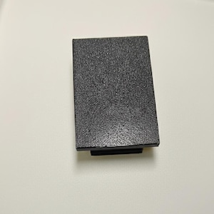 May include: A rectangular, dark gray object with a textured surface. The object has a black base and is set against a white background. The texture appears to be a speckled or granular pattern.
