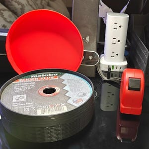 May include: A stack of metal cutting discs with the text "Metabo" and "Turbo Plus". A red bowl and a red tape measure are also visible. A white power strip with multiple outlets and USB ports is in the background.