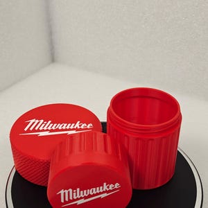 May include: Three red containers with the Milwaukee logo in white. One is cylindrical with a ridged exterior, another is a lid with a ridged edge, and the third is a flat lid. The containers are on a black and white surface.