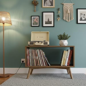 Puede incluir: Un salón de estilo moderno de mediados de siglo con un mueble tocadiscos de madera, una lámpara tejida y obras de arte enmarcadas. El mueble contiene un tocadiscos, discos de vinilo y libros. Las paredes están pintadas de color turquesa y el suelo tiene una alfombra gris.