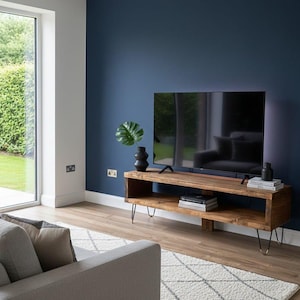 May include: A wooden TV stand with a rectangular shape and hairpin legs. The stand is made of light brown wood and has an open shelf. A large black television sits on top, with a dark blue wall in the background.