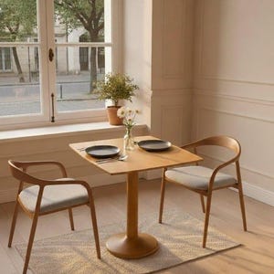 Könnte beinhalten: Ein quadratischer Holztisch mit Mittelfuß und zwei passenden Holzstühlen mit hellgrau gepolsterten Sitzflächen. Der Tisch ist mit zwei dunklen Tellern und einer klaren Glasvase mit weißen Blumen gedeckt. Eine kleine Topfpflanze steht auf dem Fensterbrett.