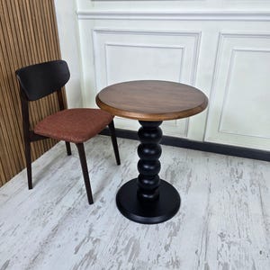 Könnte beinhalten: Ein kleiner, runder Holztisch mit einer dunkelbraunen Tischplatte und einem schwarzen, kugelgestapelten Fuß. Neben dem Tisch steht ein Holzstuhl mit einem rotbraunen Sitz und einer dunkelbraunen Rückenlehne. Der Tisch hat einen Durchmesser von etwa 61 cm.