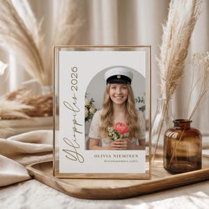 May include: A framed photograph featuring a young woman holding a pink flower, wearing a white cap. The photo is in a gold frame, with text that reads "Ylioppilas 2026" and the name "Olivia Nieminen". The background includes dried plants and a vase.