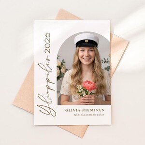May include: A graduation announcement card featuring a smiling person holding a pink flower. The card has the text "Ylioppilas 2026" in gold script, along with a name and school. The card is on a peach-colored envelope.