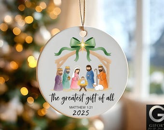 May include: A round Christmas ornament with a Nativity scene illustration. The ornament features a green bow, a stable, and figures of the Holy Family and the Three Wise Men. Text reads "the greatest gift of all" and "Matthew 1:21 2025".