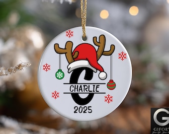 May include: A white ceramic Christmas ornament with a reindeer design. The ornament features a red Santa hat, brown antlers, and the name "Charlie" with the year "2025". Green and red ornaments hang from the antlers, and red snowflakes are scattered around the edge.