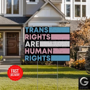 Może przedstawiać: Czarny znak ogrodowy z napisem "TRANS RIGHTS ARE HUMAN RIGHTS" w kolorze niebieskim i różowym, z projektem flagi transgenderycznej. Znak jest wyświetlany na zielonym trawniku przed domem.