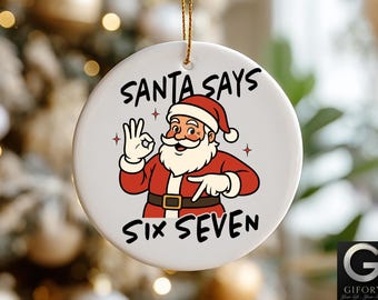 May include: A white ceramic Christmas ornament with a cartoon Santa Claus illustration. The text "SANTA SAYS SIX SEVEN" is printed in black. Santa is wearing a red suit and hat, with a white beard and gloves.