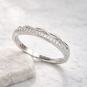 Peut inclure: Une bague argentée avec un motif tressé et une rangée de petites pierres claires. La bague est posée sur une pierre blanche sur fond blanc. La bande de la bague mesure environ 2 mm de large.