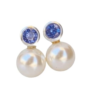 May include: A pair of elegant earrings featuring a large, lustrous white pearl base topped with a round, blue gemstone set in a silver-colored bezel. The earrings have a classic, sophisticated design.
