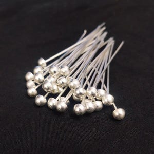 May include: A collection of silver-colored head pins with a small, round bead at the top of each pin. The pins are arranged in a loose pile against a dark background, showcasing their reflective quality. These pins are likely used for jewelry making or crafting.