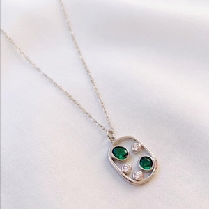 May include: A silver necklace with an oval pendant. The pendant features two oval-cut emerald green gemstones and two smaller, round, clear gemstones. The necklace is displayed on a white, textured surface.
