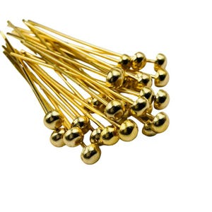 May include: A collection of gold-colored head pins with rounded, ball-shaped heads. The pins are slender and have a shiny finish, ideal for jewelry making or craft projects. The pins are scattered against a white background.