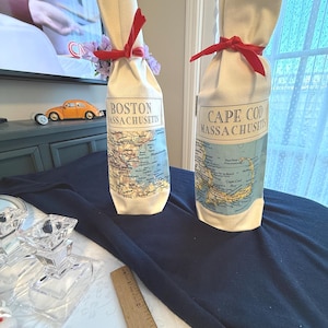 May include: Two cream-colored wine bottle bags with red ribbons. One bag has "Boston Massachusetts" printed on it, and the other says "Cape Cod Massachusetts". Both bags feature a map design.