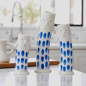 Op de afbeelding: Drie witte keramische vazen met blauwe ovale patronen. Elke vaas heeft een gezicht met kleine zwarte stippen voor ogen en een neus. De vazen hebben kleine armachtige uitsteeksels. De vazen staan op een wit aanrecht.