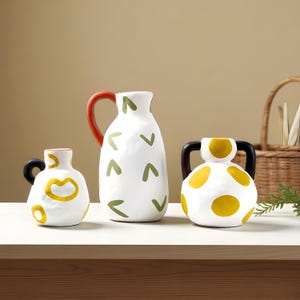 May include: Three white ceramic vases with colourful designs. The vase on the left has a black handle and yellow accents. The middle vase has a red handle and green accents. The vase on the right has black handles and yellow polka dots.