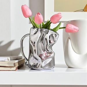 May include: A silver-coloured vase with a handle, containing pink tulips with green stems and leaves. The vase has a distinctive, crumpled design. A white ceramic vase with a pink tulip is also visible.