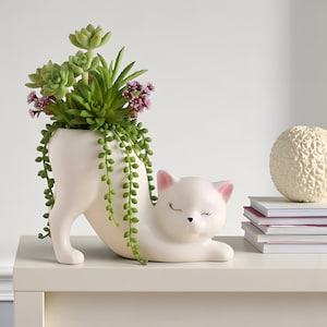 Könnte beinhalten: Ein weißer Keramik-Katzen-Pflanztopf mit Sukkulenten. Die Katze liegt entspannt, die Augen geschlossen. Der Topf enthält grüne Sukkulenten und kleine lila Blüten.