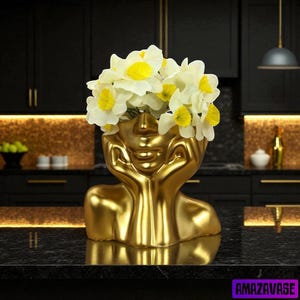 May include: A gold-colored decorative vase shaped like a human head, holding white and yellow flowers. The vase has a smooth, reflective surface and is resting on a black countertop. The flowers are white with yellow centers.