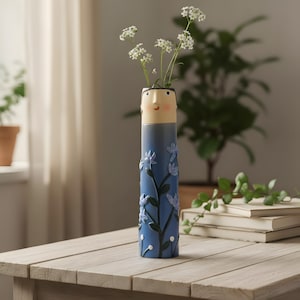 Pode incluir: Um vaso de cerâmica azul com um design de rosto no topo, segurando flores brancas. O vaso apresenta um padrão floral em relevo em um azul mais claro. Está em uma mesa de madeira.