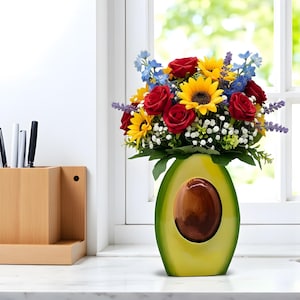 May include: A vibrant floral arrangement in an avocado-shaped vase. The bouquet features red roses, yellow sunflowers, blue delphiniums, and sprigs of lavender. The ceramic vase is green with a brown pit-shaped opening, set on a white countertop.