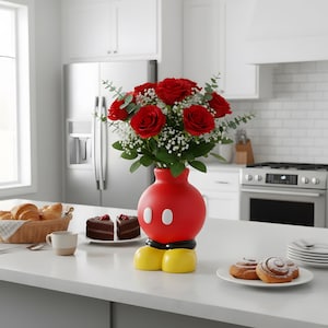 Könnte beinhalten: Eine rote Vase in Form eines Zeichentrickkopfes mit weißen Knöpfen und gelben Füßen hält einen Strauß roter Rosen und weißer Füllblumen. Die Vase steht auf einer weißen Arbeitsplatte in einer Küche.