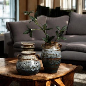 May include: Two ceramic vases with handles, one holding green leafy branches, sit on a wooden table. The vases have a textured, layered design in shades of brown, white, and blue. A stack of small ceramic pieces is visible in the background.