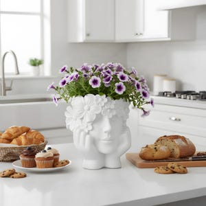 Puede incluir: Una maceta blanca de cerámica con forma de rostro y corona floral contiene petunias moradas y blancas. Productos horneados, incluyendo cruasanes, magdalenas y pan, están dispuestos sobre una encimera blanca en una cocina.