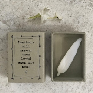 Può includere: Una piccola scatola rettangolare con la scritta "Feathers will appear when loved ones are near" e una piuma bianca all'interno. La scatola è aperta, rivelando la piuma. Un fiore bianco è sopra la scatola.