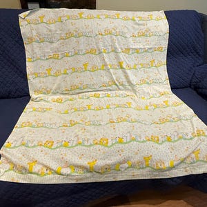 Peut inclure: Une couverture pour bébé à motifs blancs et jaunes avec des rangées d'animaux de dessins animés, dont des lions et des girafes, avec une ligne ondulée verte et de petites étoiles jaunes. La couverture est pliée sur un canapé bleu foncé.