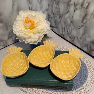 May include: Three yellow pineapple-shaped ceramic dishes with textured surfaces. The dishes are arranged on a green book, with a white flower and a marble background. The dishes are designed to resemble pineapples.