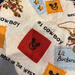 Puede incluir: Tela blanca con tem&aacute;tica de vaquero con personajes de dibujos animados, estrellas y el texto "#1 Cowboy" y "Best in the West". El dise&ntilde;o incluye parches rojos, amarillos y azules con iconos de Mickey Mouse.