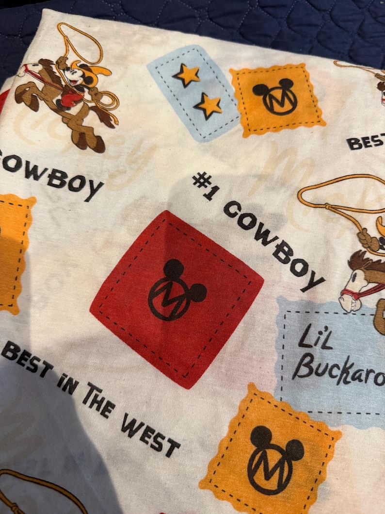 Puede incluir: Tela blanca con tem&aacute;tica de vaquero con personajes de dibujos animados, estrellas y el texto "#1 Cowboy" y "Best in the West". El dise&ntilde;o incluye parches rojos, amarillos y azules con iconos de Mickey Mouse.